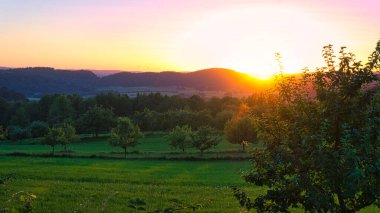 Saarland 'da bir çayırda gün batımında ağaçlar ve vadi manzarası. Sıcak hava turuncu, kırmızı ve altın renginde.