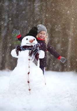 Büyük gülümseyen kardan adam, kara şapkalı, kış günü ormanın yanındaki küçük bir kızın yanında duruyor..