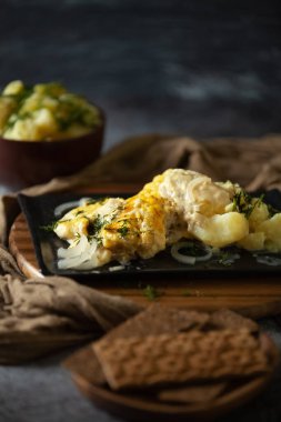 Koyu arkaplanda fırında balık ve çerez sosu olan bir güveç, kilden bir kasede patates, yanında da küçük miktarda ekmek.