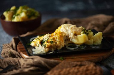 Koyu arkaplanda fırında balık ve çerez sosu olan bir güveç, kilden bir kasede patates, yanında da küçük miktarda ekmek.