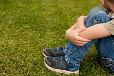 Kirli dizleri olan mavi kotlu bir çocuk, bir tişört ve mavi eklemli koyu renkli spor ayakkabılar yeşil çimlerin üzerinde oturuyor, dizlerine mesaj atmak için bir yer ile sarılıyor.