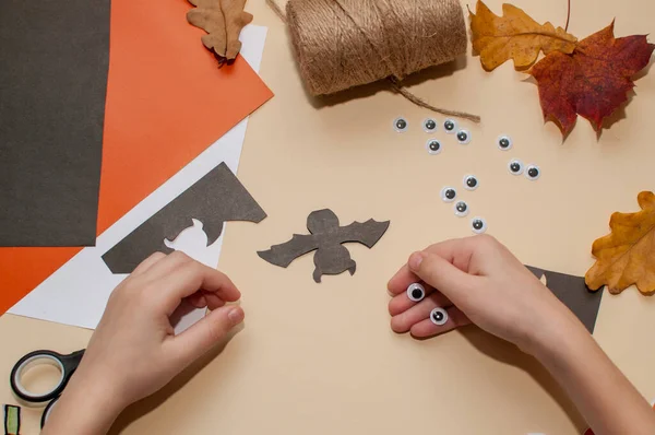 子供はおもちゃの目の横にベージュの背景に黒い紙で作られた目でバットの形でハロウィーンの工芸品を作ります ロープと乾燥葉 — ストック写真