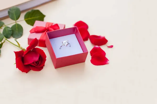 緑の茎に赤いバラの横にベージュの背景に弓を持つ開いて赤いギフトボックスに大きな宝石と白い金の指輪とテキストのための場所と赤い花弁 — ストック写真