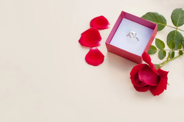 緑の茎に赤いバラの横にベージュ色の背景に開いた赤いギフトボックスに大きな宝石を持つ白い金の指輪とテキストのための場所と赤い花弁 — ストック写真