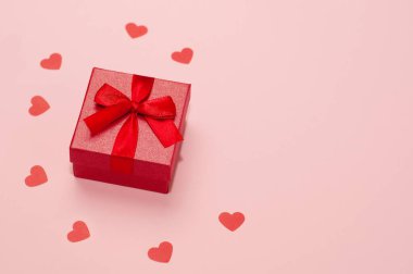 Pembe arka planda kırmızı yüzük kutusu ve kalp konfetisi.