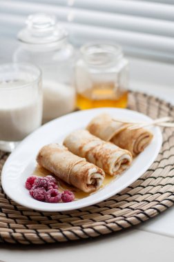 Krep, rulo şeklinde, donmuş ahududu ve bal kaplı beyaz bir tabakta bir bardak sütün arka planında ahşap çatallar, şeker kavanozları ve balla hasır peçetenin üzerinde.