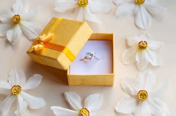ダフォディルの花の光を背景に宝石のような白い金のリングを持つ黄色の箱を開く — ストック写真