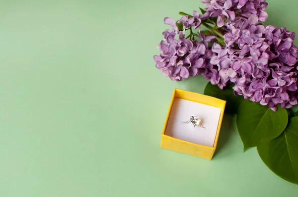 在绿色背景的紫丁香花旁的一个黄色盒子里有一块大石子的戒指 — 图库照片