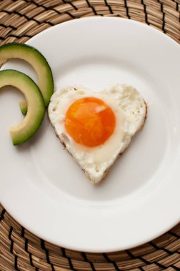 kalp şeklinde çırpılmış yumurta, beyaz bir tabakta, avokado dilimleri ve jüt peçeteyle.