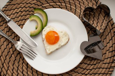 Avokado dilimleri, aletler ve pişirme kalıbıyla beyaz bir tabakta kalp şeklinde çırpılmış yumurta.