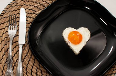 Kalp şeklinde çırpılmış yumurta, siyah bir tabakta, çatal bıçaklı bir peçetenin üzerinde.