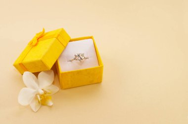 Bej arkaplanda orkide çiçeğinin yanında sarı bir kutuda büyük bir mücevher olan bir yüzük.