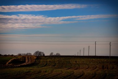 Letonya 'da sonbaharda tarımdan geçen toprak toprak yol. Elektrik direkleri ve kablolar, ilginç bulutları olan güzel mavi gökyüzü.