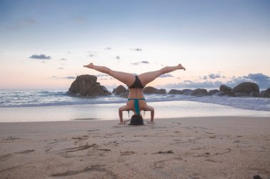 Gün batımında plajda yoga yapan mavi bikinili genç bir kadın, fitness konsepti, esmer sörfçü kız, asana amuda kalkıp bacaklarını ayıran sağlıklı aktif yaşam tarzı konsepti.. 