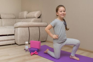 Evde lavanta halısında spor yapan gülümseyen küçük kız. Tek dizinin üstünde, elleri kemerin üstünde. Kettlebells yakınlarında, yoga blokları su şişesi.