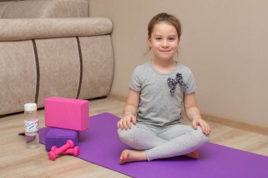 Güzel bir lavanta halıya oturmuş, yoga pozunda, nilüfer pozisyonunda gülümseyen küçük kız. Yakınlarda yoga blokları, çaydanlıklar ve bir şişe su var..