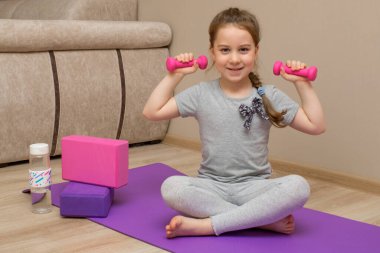 Küçük gülümseyen bir kız spor paspasına oturur ve elinde kettlebell taşır. O, evde spor yapar. Yanında yoga blokları ve bir şişe su var..