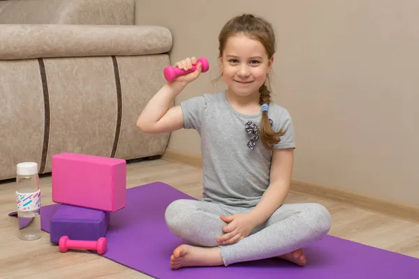 A little smiling girl sits on a fitness mat and holds a kettlebell in her right hand. She plays sports at home. Next to her are yoga blocks and a bottle of water. — Stock Photo, Image