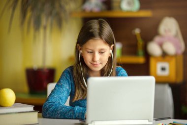 Kız kulaklıkla dizüstü bilgisayarın önünde oturuyor ve ekrana bakıyor. Evden online öğrenme