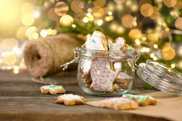 Glass jar filled with homemade cookies stands on a wooden table in the kitchen. Wrapping paper and rope are prepared for packing sweet gifts for Christmas.