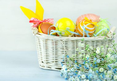 Bir sepet Paskalya yumurtası mavi ahşap bir masanın üzerinde duruyor. Bahar beyaz ve mavi çiçekler yan yana uzanır.