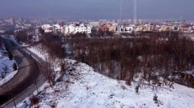 Kış boyunca şehrin kenar mahallelerinde karla kaplı evler ve küçük bir koruluk. İkisinin arasında elektrik kuleleri var. Şehir ve dağlar uzaktan görülebilir. Hava görüntüleri