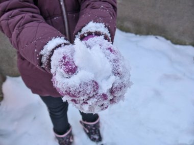 Karı tutan bir çocuğun ellerini yakın plan çek. Çocuk mor eldiven ve mor bir ceket giyiyor. Arka planda gri duvar, yerde kar.