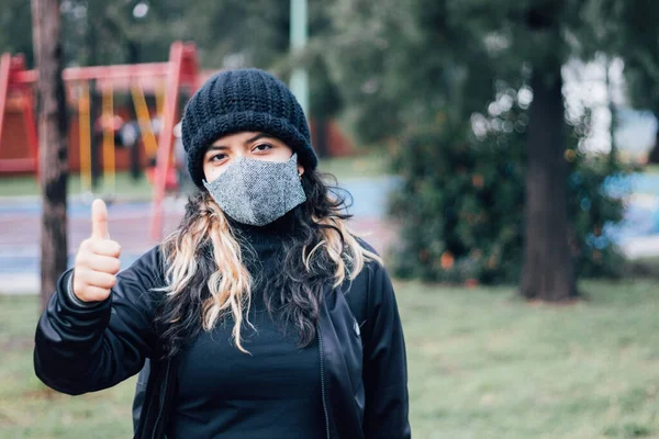 Latin üniversiteli kız, yüz maskesi takmış parkta dikilirken başparmağını kaldırdı.