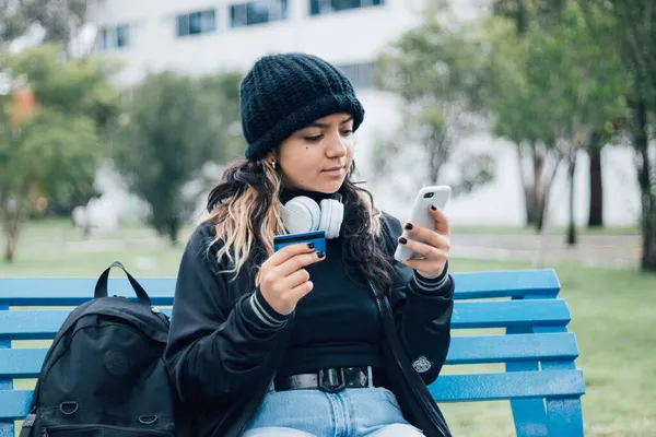 Latin üniversiteli bir kız parkta bankta oturuyor ve kredi kartıyla online alışveriş yapıyor.
