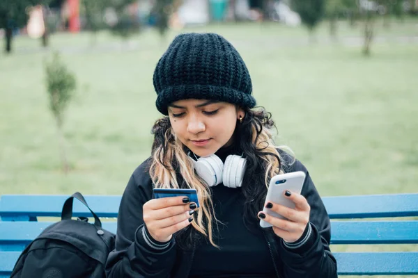 Latin üniversiteli bir kız parkta bankta oturuyor ve kredi kartıyla online alışveriş yapıyor.