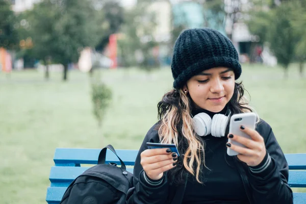 Latin üniversiteli bir kız parkta bankta oturuyor ve kredi kartıyla online alışveriş yapıyor.