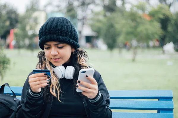 Latin üniversiteli bir kız parkta bankta oturuyor ve kredi kartıyla online alışveriş yapıyor.