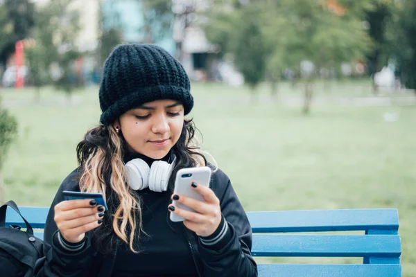 Latin üniversiteli bir kız parkta bankta oturuyor ve kredi kartıyla online alışveriş yapıyor.