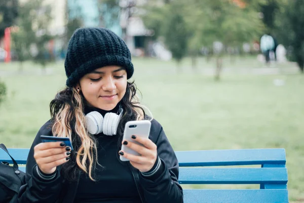Latin üniversiteli bir kız parkta bankta oturuyor ve kredi kartıyla online alışveriş yapıyor.