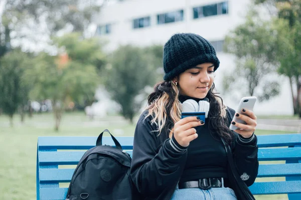 Latin üniversiteli bir kız parkta bankta oturuyor ve kredi kartıyla online alışveriş yapıyor.