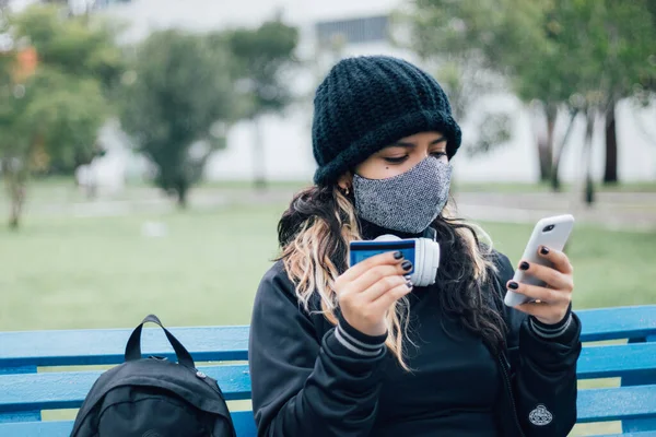 Latin üniversiteli bir kız parkta bankta oturuyor ve kredi kartı ve yüz maskesi ile online alışveriş yapıyor.