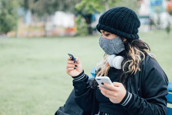 Latin üniversiteli bir kız parkta bankta oturuyor ve kredi kartı ve yüz maskesi ile online alışveriş yapıyor.