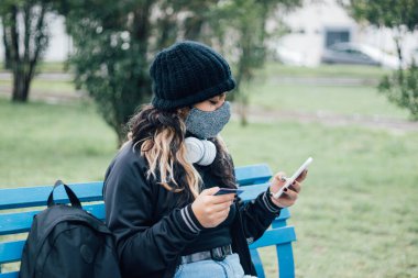 Latin üniversiteli bir kız parkta bankta oturuyor ve kredi kartı ve yüz maskesi ile online alışveriş yapıyor.