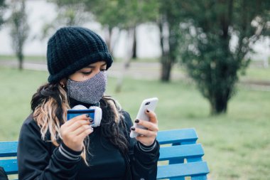 Latin üniversiteli bir kız parkta bankta oturuyor ve kredi kartı ve yüz maskesi ile online alışveriş yapıyor.