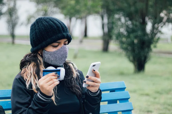 Latin üniversiteli bir kız parkta bankta oturuyor ve kredi kartı ve yüz maskesi ile online alışveriş yapıyor.