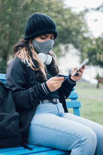 Latin üniversiteli bir kız parkta bankta oturuyor ve kredi kartı ve yüz maskesi ile online alışveriş yapıyor.