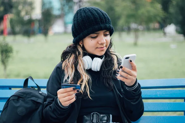 Latin üniversiteli bir kız parkta bankta oturuyor ve kredi kartıyla online alışveriş yapıyor.