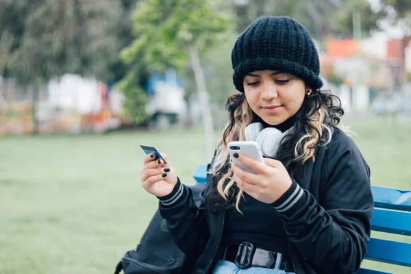 Latin üniversiteli bir kız parkta bankta oturuyor ve kredi kartıyla online alışveriş yapıyor.