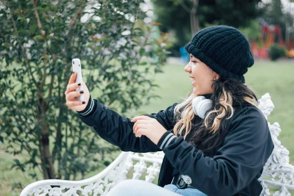 Adolescente latina universitaria sentada en un banco en el parque tomando una selfie en su teléfono —  Fotos de Stock