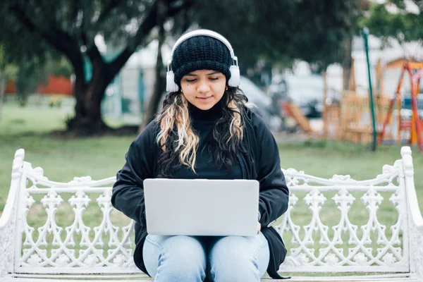 Latin üniversiteli bir kız parkta bir bankta dizüstü bilgisayar kullanarak oturuyor.