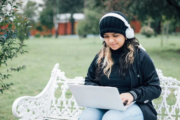 Latin üniversiteli bir kız parkta bir bankta dizüstü bilgisayar kullanarak oturuyor.