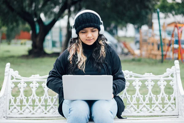 Latin üniversiteli bir kız parkta bir bankta dizüstü bilgisayar kullanarak oturuyor.