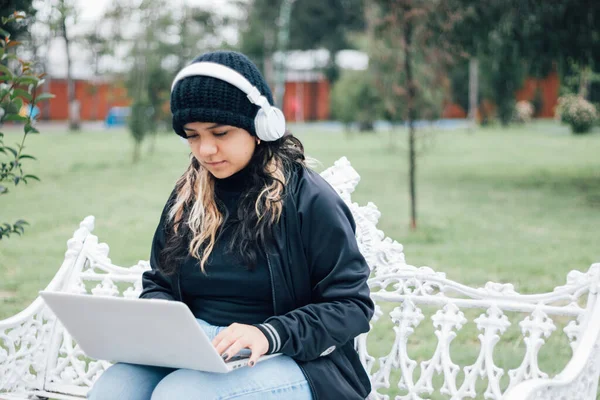 Latin üniversiteli bir kız parkta bir bankta dizüstü bilgisayar kullanarak oturuyor.