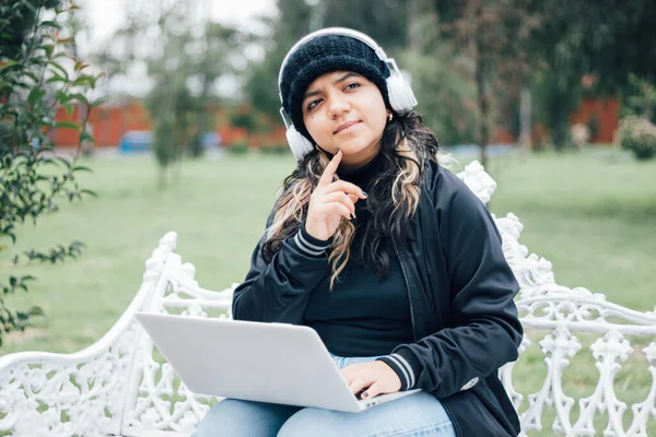 Latin üniversiteli bir kız parkta bankta oturmuş bilgisayarla düşünüyor.