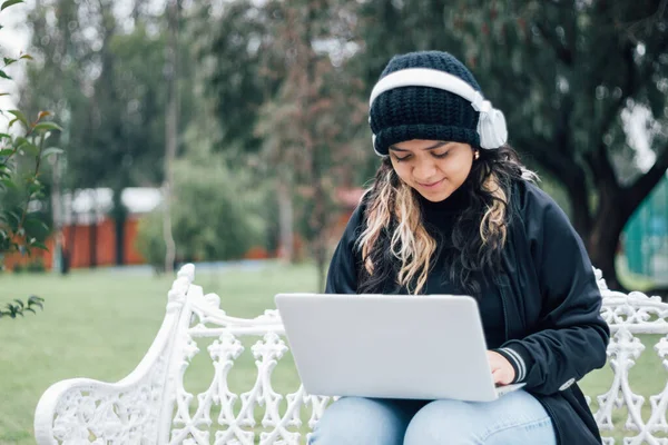 Latin üniversiteli bir kız parkta bir bankta dizüstü bilgisayar kullanarak oturuyor.